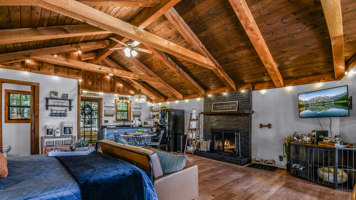 A welcoming open-plan living area featuring exposed wooden beams, a cozy fireplace, comfortable seating, and a view of the adjacent kitchen area. The space is bright and inviting, suggesting a convenient transition from arrival to relaxation.
