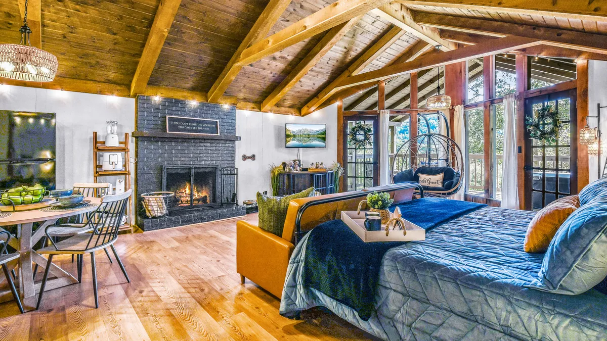 The image showcases the inviting interior of Firefly Lookout cabin, featuring a modern fireplace as the centerpiece, surrounded by large windows that flood the space with natural light. A cozy hanging chair beckons for quiet moments, while a dining table invites guests to gather and share meals. The atmosphere is bright, freshly redecorated, and designed for both relaxation and connection.