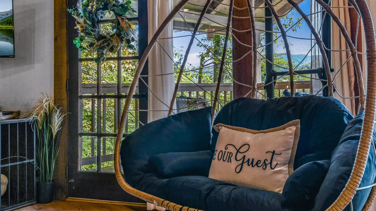 A cozy hanging chair with a decorative pillow reading 'Our Guest' is placed near a door with a view outside. Soft natural light fills the space, creating a serene atmosphere that hints at peaceful mornings or evenings spent with a pet.
