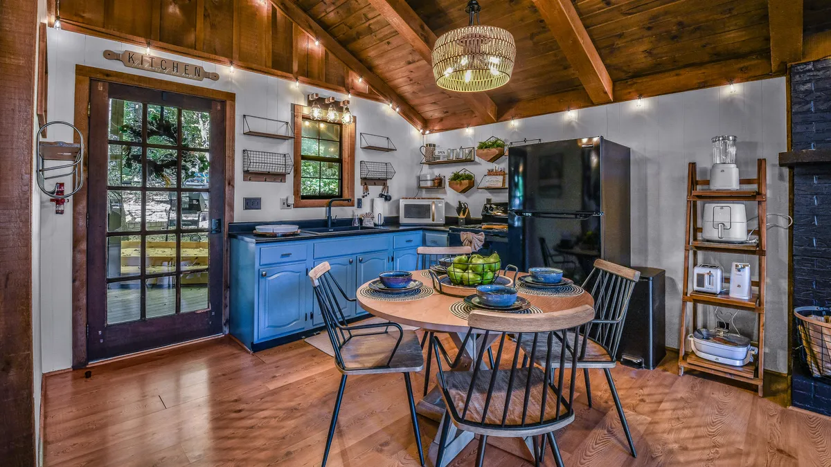 A cozy dining space with a wooden table for four, accompanied by blue cabinetry, charming lighting, and windows offering serene greenery views. The table is set for meals or board games, with an inviting, communal appeal.