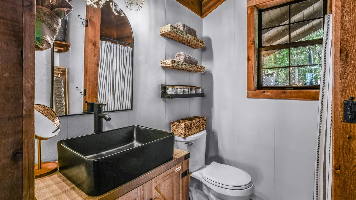 A warmly lit bathroom space featuring a black sink, wooden shelving, a decorative mirror, and plenty of natural light streaming through a window. The inviting setup hints at a tranquil spot for self-care routines, with clean towels and thoughtful design touches visible.