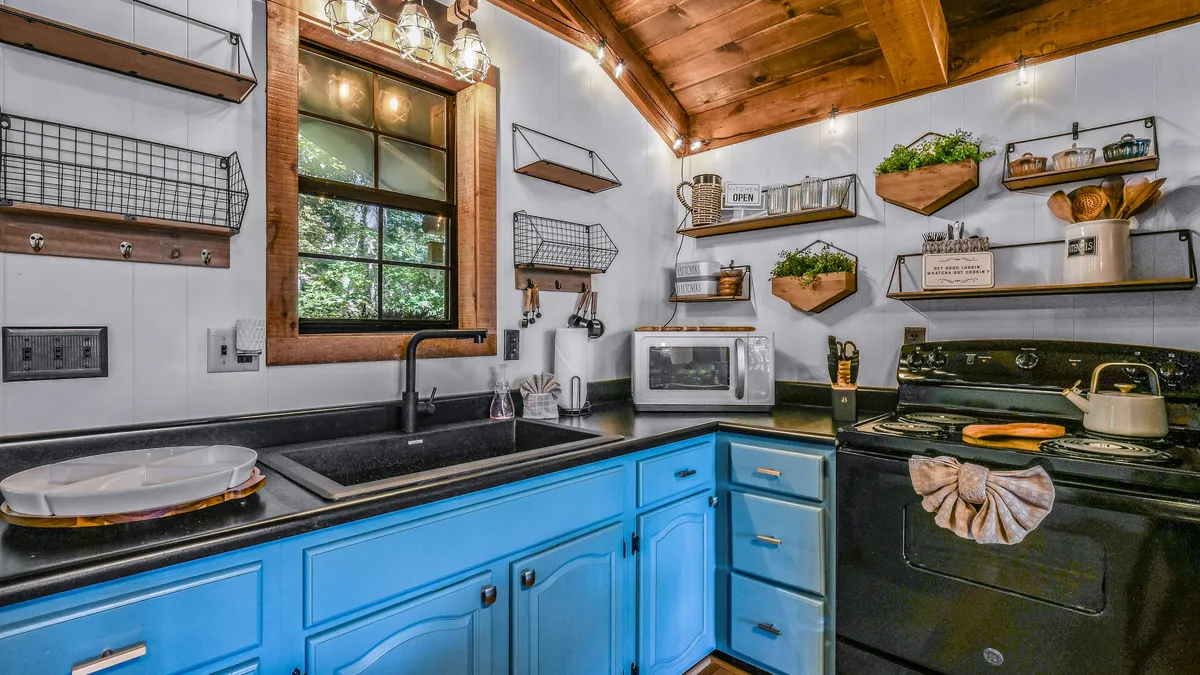 A warmly lit kitchen scene featuring blue cabinets, a sleek black countertop, window streaming in natural light, and decorative shelves displaying kitchen essentials. The layout exudes functionality and style, perfect for culinary creativity and gatherings.