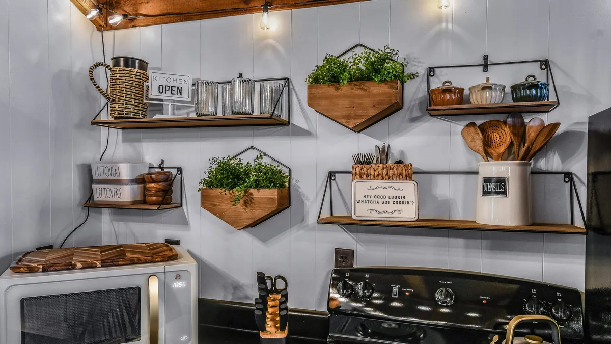 A well-stocked, open kitchen scene with wooden shelves lined with essentials, hanging plants adding a fresh touch, and an 'open kitchen' sign that welcomes guests to prepare meals or snacks at their convenience.