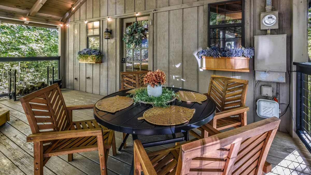 A serene dining space with a wooden table and chairs surrounded by decorative plants, basking in soft natural light. The setup hints at the transition to an outdoor deck or patio, with subtle cues of relaxation and togetherness.