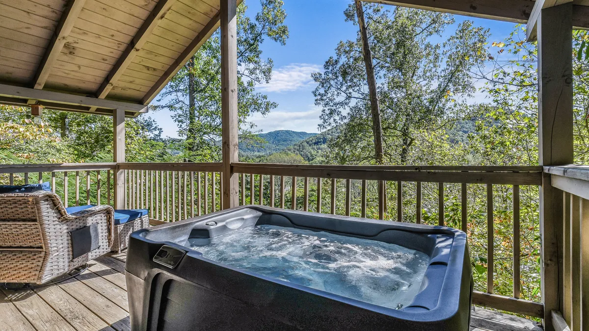 A serene outdoor wooden deck featuring a bubbling hot tub with a panoramic mountain view. A comfortable chair sits nearby, inviting guests to relax and soak in the scenery. The setting is perfect for enjoying sunsets, stargazing, or unwinding after a day of adventure.