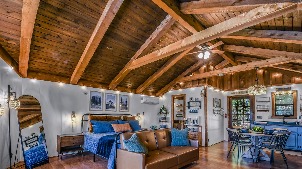 A warm, inviting open-plan studio featuring a plush sofa, wooden beams, a cozy bed with soft linens, and a dining table—capturing both the living and sleeping areas in a single, connected space. Natural light fills the room, and modern yet rustic décor accents create a comfortable, mountain-chic atmosphere.