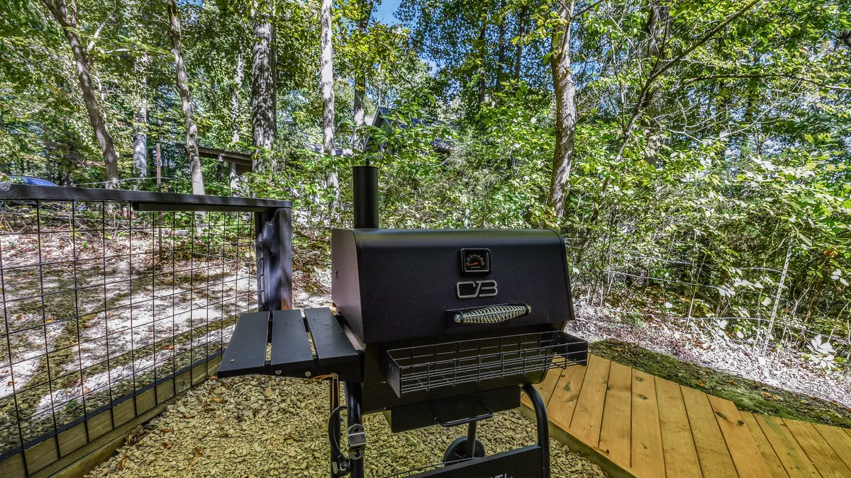 A spacious deck area with a charcoal grill set against a backdrop of lush greenery. The setup is casual yet inviting, suggesting outdoor feasts and relaxed barbecue gatherings as the sun sets.